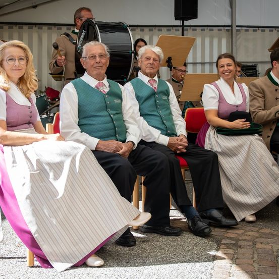 Eine Gruppe älterer Personen in traditioneller Kleidung sitzt auf Stühlen, lächelt und posiert für ein Foto, während eine Band im Hintergrund spielt.