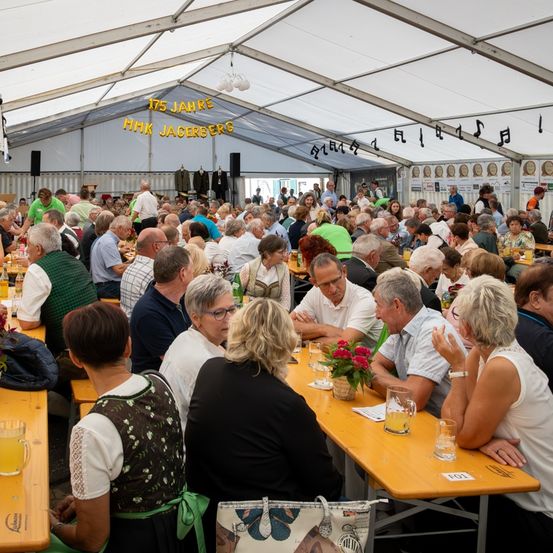 Eine große Gruppe von Menschen sitzt in einem Zeltrestaurant, einige in traditioneller Kleidung. Die Tische sind mit Essen und Getränken gefüllt. Ein Banner lautet '175 Jahre MHK Jaceberg'. Über den Tischen hängen Musiknoten.