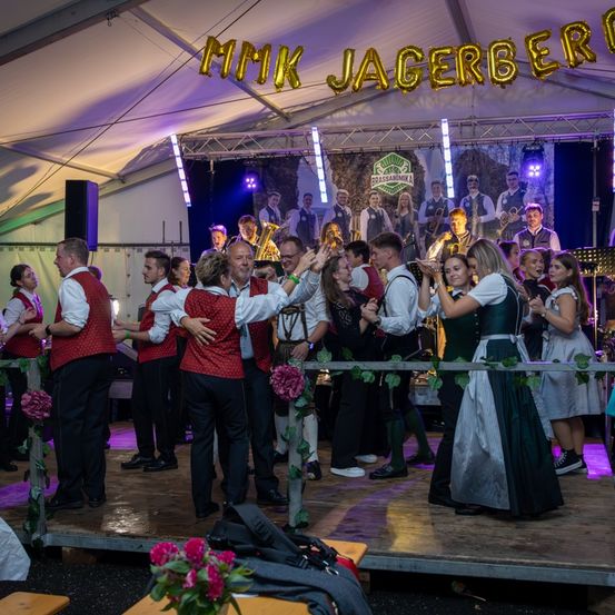 Eine Gruppe von Menschen auf der Bühne in traditioneller deutscher Kleidung tanzt unter einem Banner, das 'MMK JAGERBERI' steht. Sie werden von lila Lichtern beleuchtet, mit einer Band, die hinter ihnen spielt.