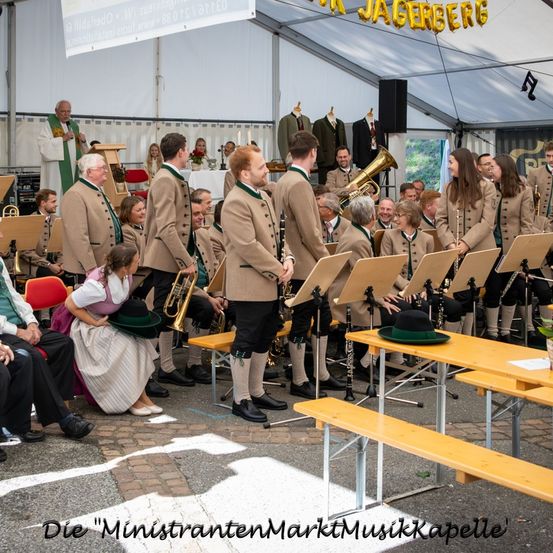Ein Musikensemble, das auf einer Bühne unter einem Zelt auftritt, mit mehreren Zuschauern in traditioneller Kleidung, einer trägt einen grünen Hut.