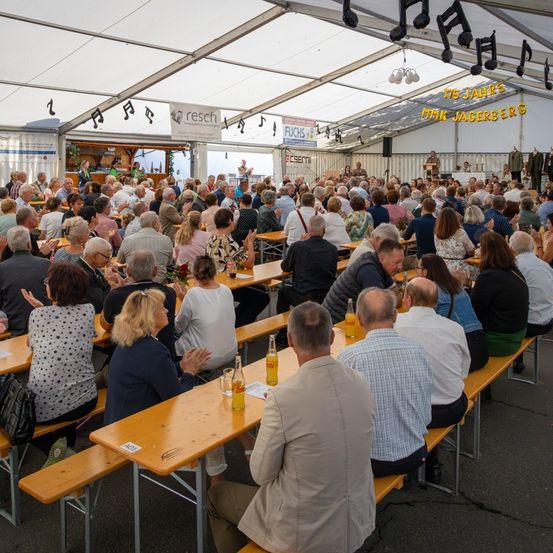 Eine große Menschenmenge versammelt sich in einem Zelt, sitzt an langen Holztischen, einige klatschen. Es hängen Noten an der Decke und ein Banner an der Wand.