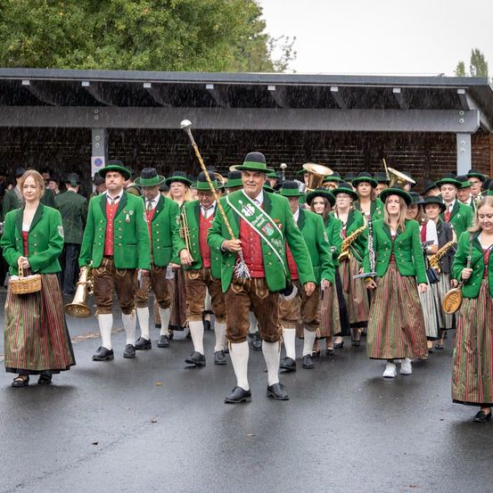 Eine Gruppe von Menschen in traditioneller bayerischer Kleidung marschiert bei Regen und hält Musikinstrumente in den Händen.