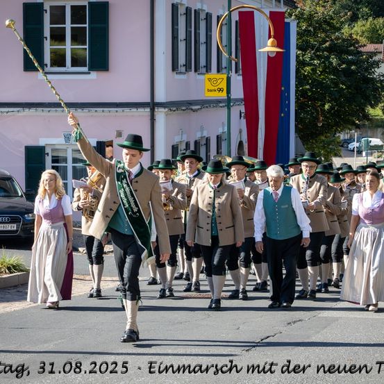 Ein Umzug mit einer Gruppe von Menschen in traditioneller bayerischer Kleidung, die auf der Straße marschieren, angeführt von einem Mann mit einem Stab.