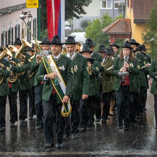 Eine Blaskapelle in grünen Uniformen zieht durch eine regennasse Straße. Sie spielen Blech- und Holzblasinstrumente. Ein Banner mit Text wird von einem Mann vorne gehalten.