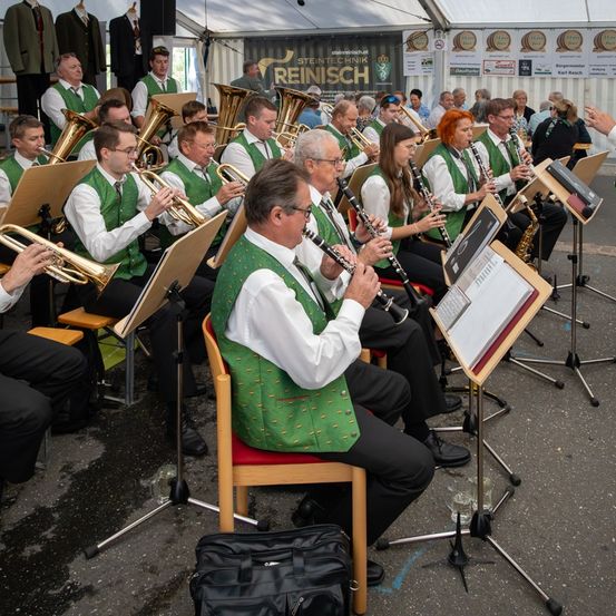 Eine Band, gekleidet in grünen Westen und weißen Hemden, tritt auf der Bühne auf. Einige spielen Trompeten, während andere Klarinetten spielen. Ein Mann im Stuhl spielt ebenfalls Klarinette. Sie tragen alle Brillen und Schuhe. Es gibt Notenständer und Taschen auf dem Boden.