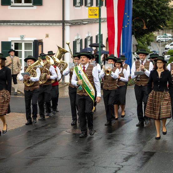 Ein Musikzug läuft auf einer nassen Straße, trägt Instrumente und trägt Uniformen. Zwei Flaggen sind sichtbar, eine ist europäisch und die andere ist österreichisch. Hinter ihnen stehen Menschen, einige halten Regenschirme. Im Hintergrund sind Gebäude, Bäume und geparkte Autos zu sehen.