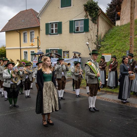 Ein Blasorchester in traditioneller deutscher Kleidung führt bei einer Parade auf, mit Zuschauern, die auf einem Hang stehen.
