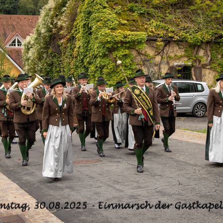 Ein Blasorchester in traditioneller bayerischer Kleidung tritt am 30.08.2025 im Freien auf. Sie spielen Instrumente, während sie eine Straße entlanggehen, mit einem Haus und einem Auto im Hintergrund.