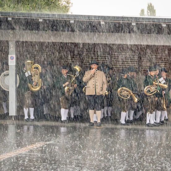 Eine Gruppe von Musikern in traditioneller bayerischer Kleidung steht in einer Reihe auf einer nassen Straße und spielt Blechblasinstrumente an einem regnerischen Tag.
