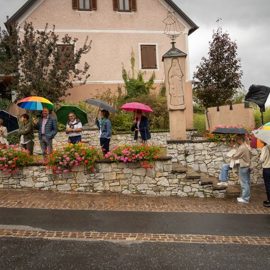 Eine Gruppe von Menschen steht unter Regenschirmen auf einem Bürgersteig bei Regen. Eine Statue und Blumen sind in der Nähe.