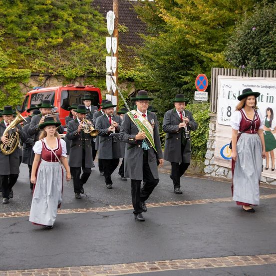Eine Blaskapelle mit Blech- und Holzblasinstrumenten spielt bei einer Parade, mit einem Feuerwehrfahrzeug im Hintergrund.