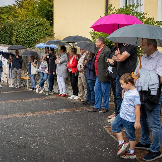 Menschen stehen in einer Reihe vor einem Gebäude, einige halten Regenschirme. Der Boden ist mit Ziegelsteinen gepflastert und das Gebiet ist von einer Steinmauer umgeben. Bäume und Pflanzen sind im Hintergrund zu sehen.