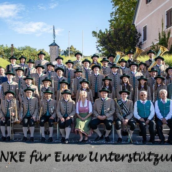 Eine Gruppe von Menschen in traditionellen Kostümen, die Musikinstrumente halten, posiert für ein Foto im Freien. Sie sitzen auf einer Steinbank mit einem Gebäude und Bäumen im Hintergrund. Ein Text-Overlay lautet: 'Danke für Ihre Unterstützung.'