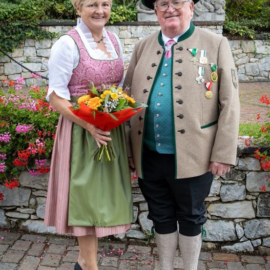 Ein Paar in traditioneller deutscher Kleidung steht auf einem Kopfsteinpflasterweg mit Blumen und einer Steinmauer im Hintergrund.