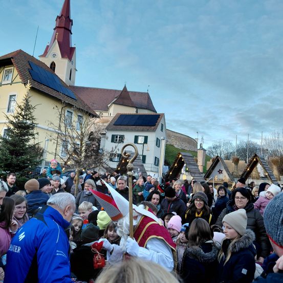 Eine Menschenmenge versammelt sich vor einer Kirche mit Solarmodulen, während eines festlichen Ereignisses mit Menschen in verschiedenen Winterkleidungen.