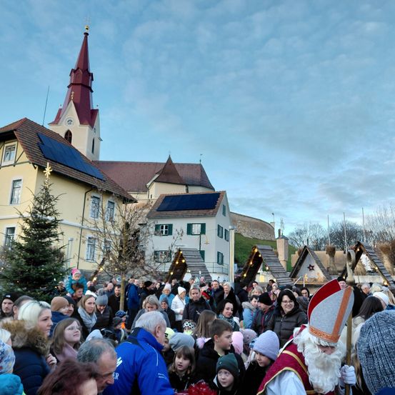 Menschenmengen versammeln sich vor einer Kirche, geschmückt mit Weihnachtsdekorationen. Eine Person, die als Weihnachtsmann verkleidet ist, steht unter ihnen und hält einen Stab. Solarmodule sind auf dem Dach sichtbar.