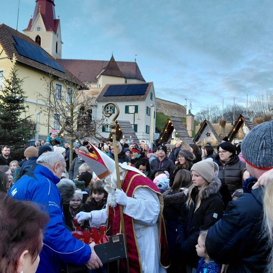 Eine Menschenmenge versammelt sich um einen als Bischof gekleideten Mann, der einen Stab hält und rote Gegenstände in einer festlichen Umgebung in der Nähe einer Kirche mit Solarmodulen verteilt.