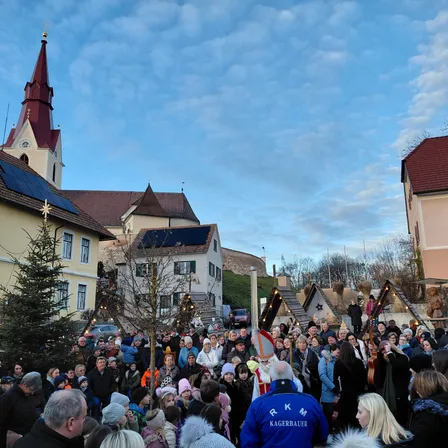 Eine Menschenmenge versammelt sich auf einem Stadtplatz mit einer Kirche und Häusern im Hintergrund. Ein Mann in einer weißen Robe steht vor einem Weihnachtsbaum, und ein Mann in einem blauen Jackett mit den Buchstaben RKM steht in der Nähe.