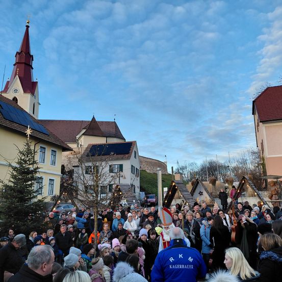 Eine Menschenmenge versammelt sich auf einem Stadtplatz mit einer Kirche und Häusern im Hintergrund. Ein Mann in einer weißen Robe steht vor einem Weihnachtsbaum, und ein Mann in einem blauen Jackett mit den Buchstaben RKM steht in der Nähe.