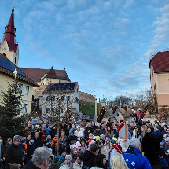 Eine Menschenmenge versammelt sich an einem bewölkten Tag vor einer Kirche. Die Kirche hat einen Turm und Solarmodule. In der Nähe befinden sich Gebäude mit roten Dächern. Eine Person in einer Bischofsmütze steht unter der Menge.