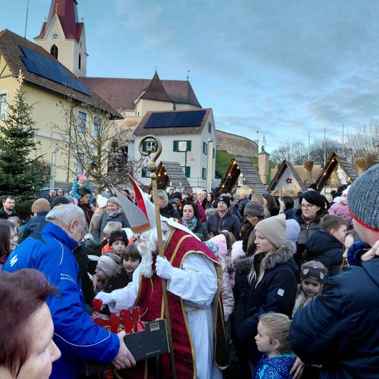 Eine große Menschenmenge versammelt sich auf einem Dorfplatz für eine festliche Veranstaltung. Ein Mann, verkleidet als St. Nikolaus, verteilt Geschenke an Kinder, während andere zusehen. Im Hintergrund steht eine Kirche mit Solarpanelen und festlichen Dekorationen.