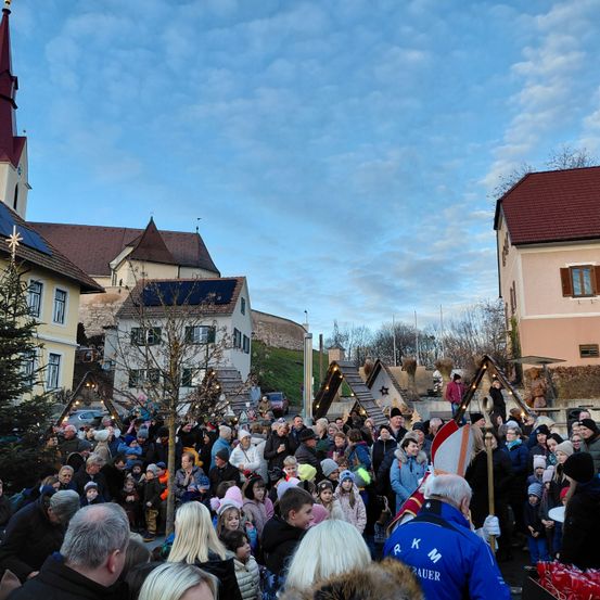 Eine Menschenmenge versammelt sich im Freien, möglicherweise für ein Festival, unter einem teilweise bewölkten Himmel. Es gibt Gebäude, Bäume und festliche Dekorationen in der Gegend. Einige Menschen tragen Mützen und Schuhe.