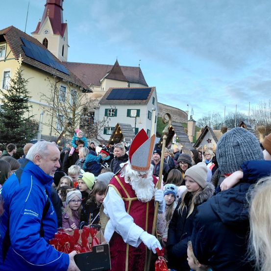 Eine Menschenmenge versammelt sich um einen als St. Nikolaus verkleideten Mann, der vor einem festlich geschmückten Stadtplatz Geschenke verteilt.