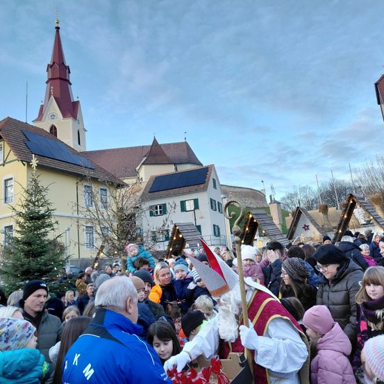 Eine große Menschenmenge versammelt sich vor einer Kirche, einige in Winterkleidung. Ein Mann als Weihnachtsmann verteilt Geschenke an die Kinder.