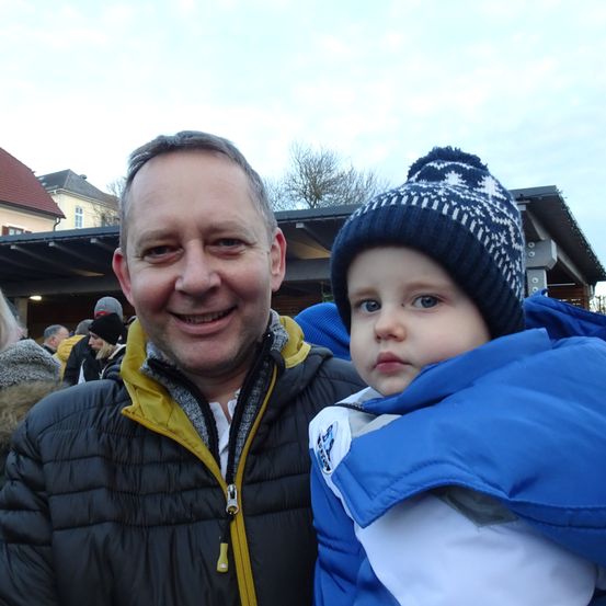 Ein Mann mit einem gelb gefütterten schwarzen Jackett und einem lächelnden Gesicht steht mit einem Kind in einem blauen Jackett. Dahinter stehen mehrere Personen im Freien.