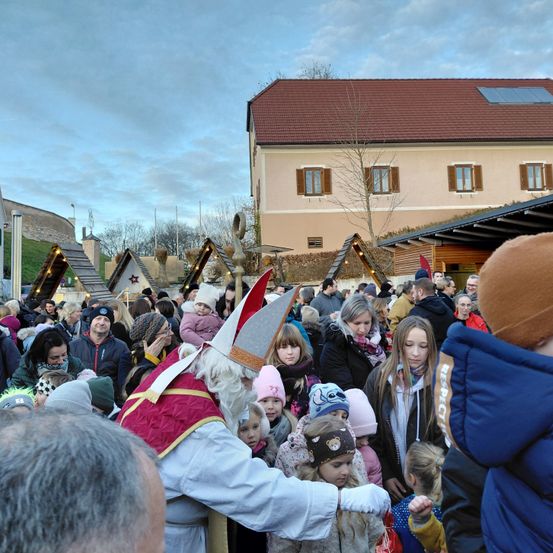 Eine Menschenmenge, darunter auch Kinder, versammelt sich um eine als Bischof verkleidete Person an einem festlichen Ort im Freien. Ein Gebäude mit Solarpanels auf dem Dach ist im Hintergrund zu sehen.