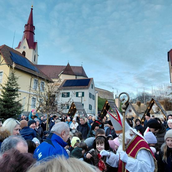 Eine Menschenmenge versammelt sich vor einer Kirche mit einem Turm, der mit Solarpanelen ausgestattet ist. Eine Person, die als Weihnachtsmann verkleidet ist, hält einen Stab und interagiert mit Kindern im Vordergrund.