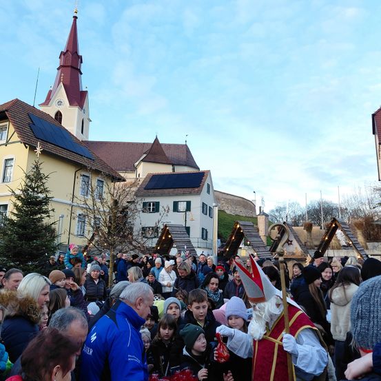 Eine Menschenmenge, darunter auch Kinder, versammelt sich vor einer Kirche, wobei ein Mann als Sankt Nikolaus verkleidet Geschenke verteilt.