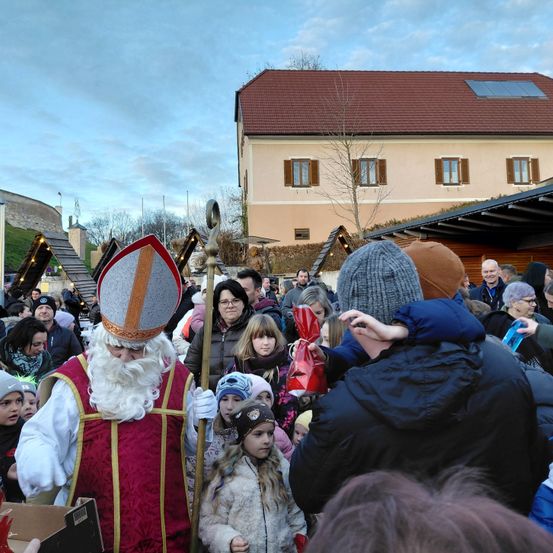 Eine Menschenmenge hat sich um eine als Sankt Nikolaus verkleidete Person versammelt, die eine rote Tasche hält, während andere zuschauen.