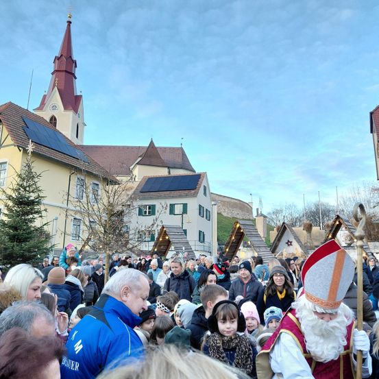Eine große Menschenmenge versammelt sich vor einer Kirche mit Solarpanelen auf dem Dach. Ein Mann, der als Sankt Nikolaus verkleidet ist, geht unter ihnen umher.