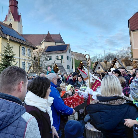 Eine Menschenmenge versammelt sich vor einer Kirche, wo ein Mann als St. Nikolaus verkleidet Geschenke an Kinder verteilt. Eine festliche Atmosphäre herrscht.