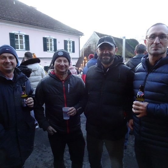 Vier Männer in Winterkleidung lächeln für ein Foto im Freien und halten Bierflaschen in den Händen. Hinter ihnen steht ein rosafarbenes Haus mit Fenstern mit Jalousien und einer Menschenmenge.