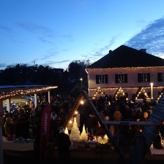 Bei Einbruch der Dunkelheit versammelt sich eine große Menschenmenge bei einer Outdoor-Veranstaltung, die von Lichterketten beleuchtet wird. Die Menge befindet sich unter einem Vordach mit Weihnachtsbäumen und anderen Dekorationen. Im Hintergrund steht ein Gebäude mit schwarzem Dach, das mit Lichtern geschmückt ist.