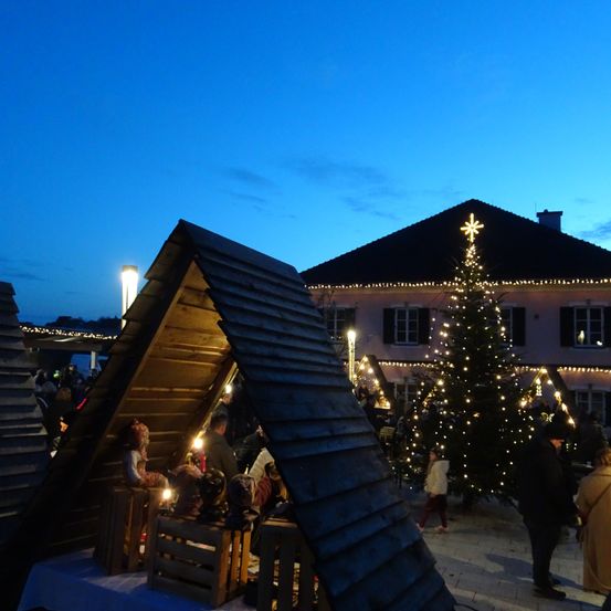 Ein Abendbild eines festlichen Marktes mit einem Weihnachtsbaum und mehreren Menschen, die herumlaufen, vor einem Haus mit beleuchteten Fenstern.