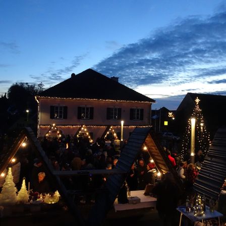 Ein festlicher Zusammenkommen unter einem Abendhimmel. Viele Menschen genießen die Nacht rund um dekorierte Zelte und Gebäude, die von Weihnachtslichtern erleuchtet sind.