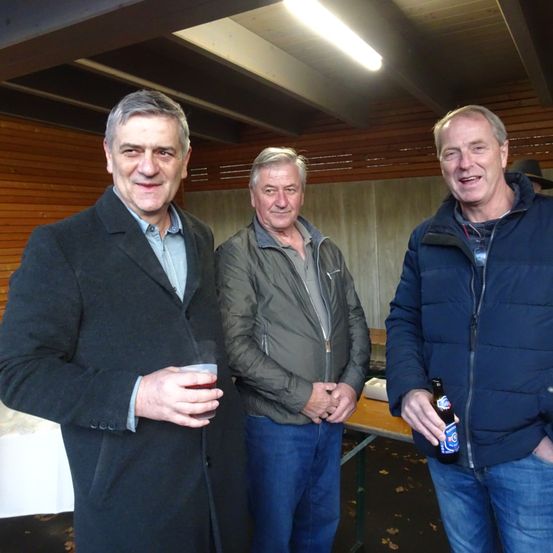 Drei Männer stehen in einem Raum mit einer Holzwand, einer hält ein Getränk, ein anderer eine Bierflasche, und ein Tisch mit einer weißen Tischdecke steht hinter ihnen.