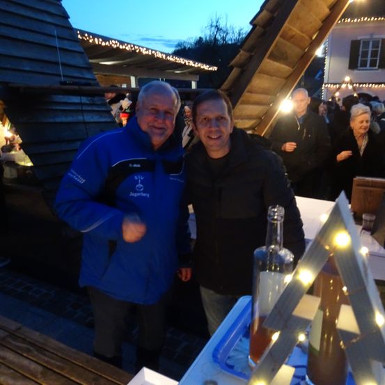 Zwei Männer stehen lächelnd bei einer Outdoor-Veranstaltung bei Nacht, mit einer hölzernen Struktur und beleuchteten Dekorationen hinter ihnen.