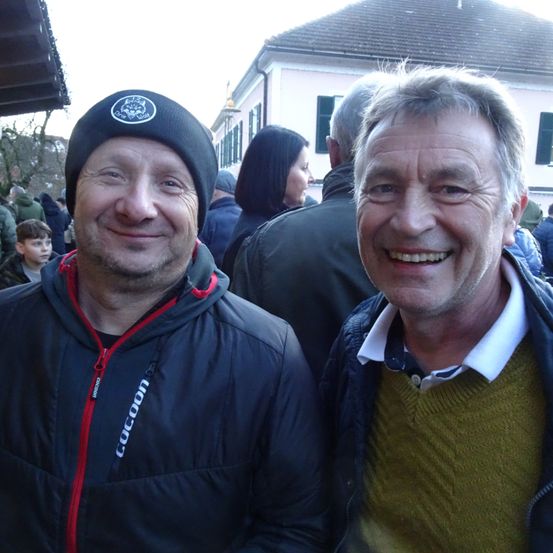 Zwei Männer lächeln für ein Foto vor einer Menschenmenge. Der Mann auf der linken Seite trägt eine Mütze mit einem Logo. Der Mann auf der rechten Seite trägt einen grünen Pullover. Hinter ihnen stehen mehrere Personen und ein Gebäude.