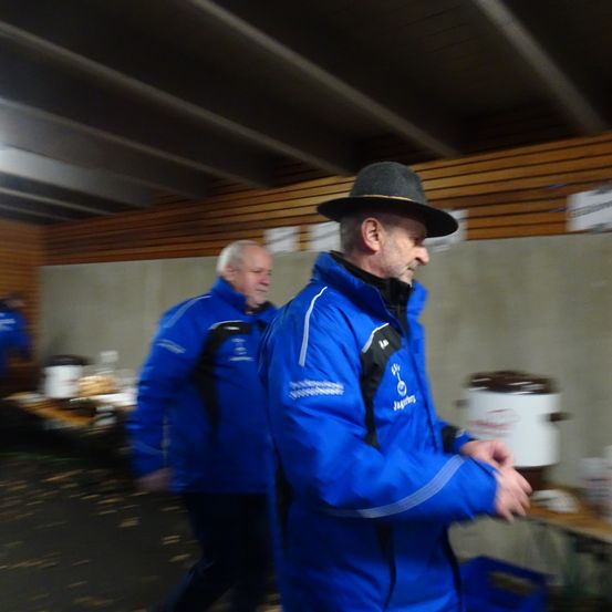 Zwei Männer in blauen Jacken, einer mit Hut, gehen durch einen Raum mit Holzwänden. Ein Mann hat einen Rucksack. Sie stehen vor einem Tisch mit einem Topf.