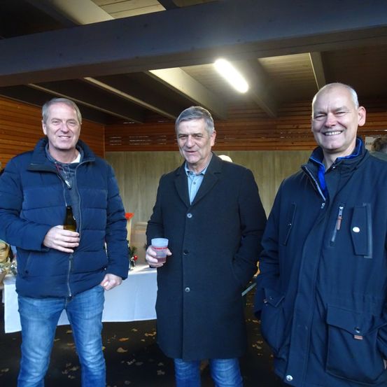 Drei Männer stehen in einem Raum zusammen. Der Mann auf der linken Seite hält eine Flasche. Der Mann in der Mitte hält einen Becher. Sie tragen alle Jacken.