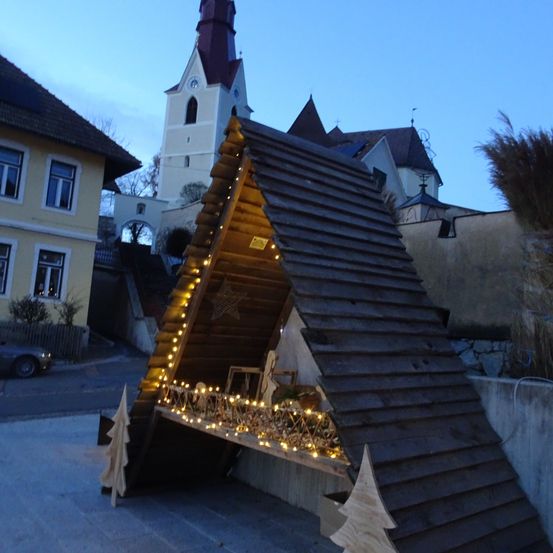 Eine beleuchtete Weihnachtskrippe steht vor einem Gebäude mit Turm.
