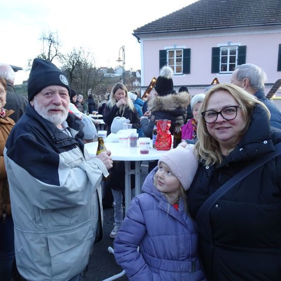 Eine Gruppe von Menschen, darunter ein älterer Mann, eine Frau und ein junges Mädchen, versammelt sich im Freien. Sie stehen in der Nähe eines Tisches mit verschiedenen Gegenständen darauf. Das Wetter ist kalt, wie ihre Winterkleidung zeigt.