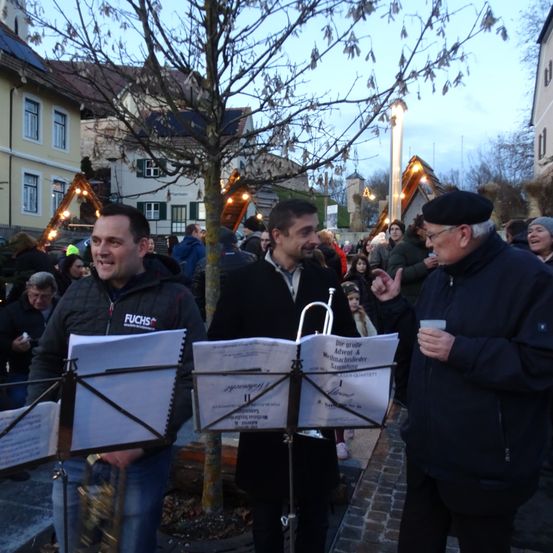 Drei Männer stehen mit Notenblättern auf einem Ständer draußen auf einem Platz, einer hält eine Trompete. Dahinter versammelt sich eine Menschenmenge auf der Straße.