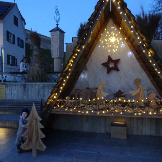 Eine dreieckige Weihnachtskrippe wird draußen beleuchtet. Ein Kind hält einen Holzbaum, während ein Stern die Wand schmückt.