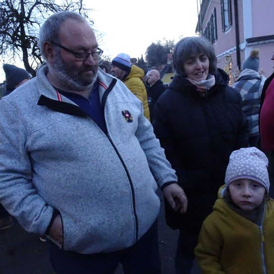 Eine Gruppe von Menschen, darunter ein Mann mit einer grauen Jacke, eine Frau mit einer schwarzen Jacke und ein Kind mit einer gelben Jacke, stehen draußen in der Nähe eines Gebäudes.