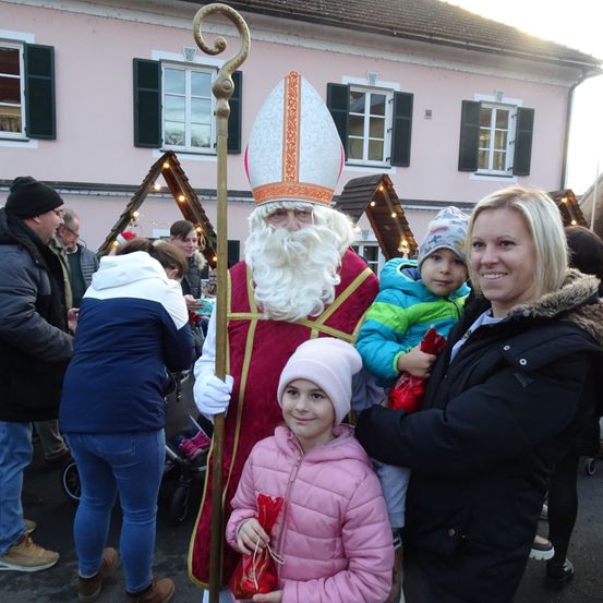 Ein Mann in der Kleidung des Heiligen Nikolaus steht mit zwei Kindern und lächelt für ein Foto. Sie stehen vor einem festlich dekorierten rosa Haus.